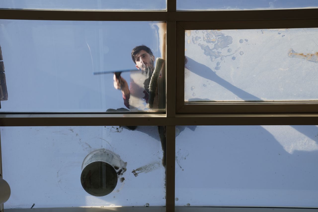 In daylight, a male window cleaner washes the exterior glass roof from above using a tool.